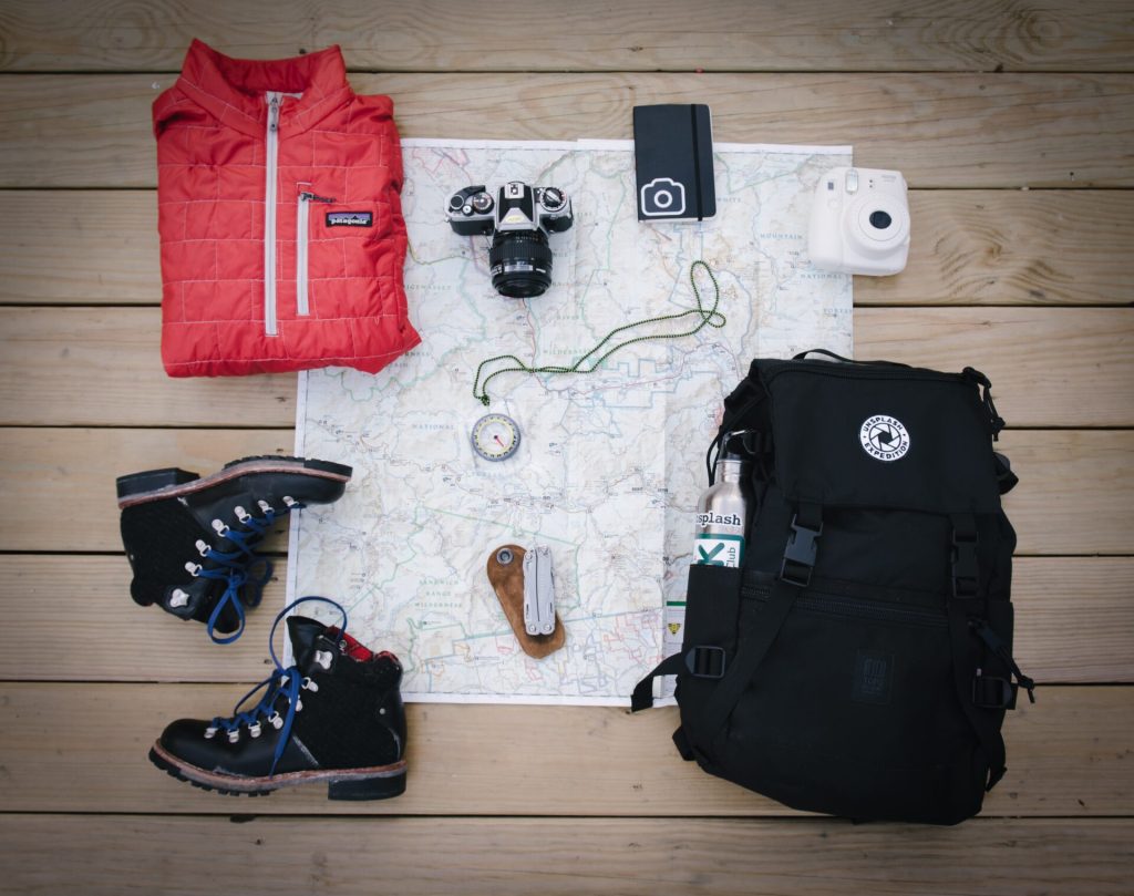 Hiking equipment laying on the floor, including a backpack, boots, a compass and a camera.