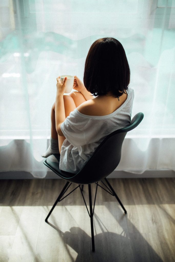 Expat Woman with black hair sitting in front of a window, looking outside and feeling lonely.