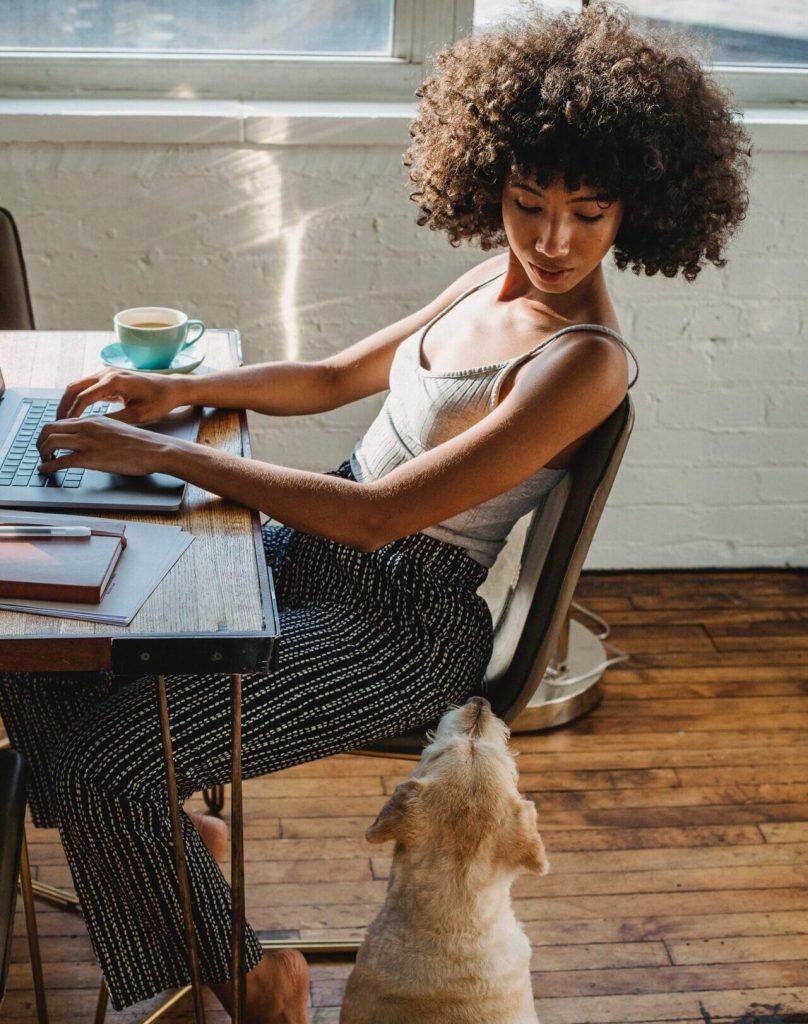 Black women sitting on a desk with a laptop looking at her dog sitting next to her on the floor.Time Management