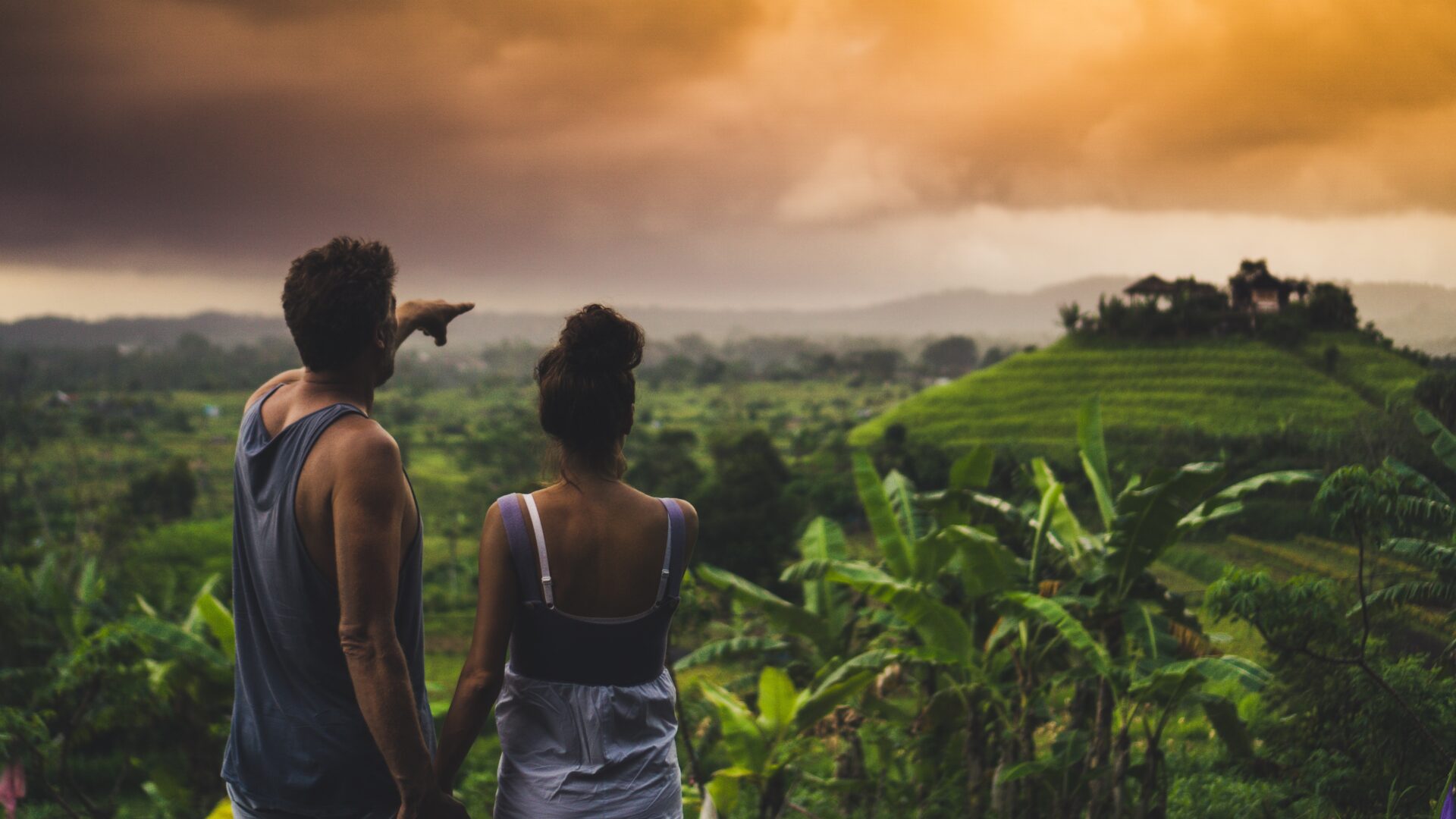 young couple from behind standing in front of a green valley in Bali, he is pointing to something far away