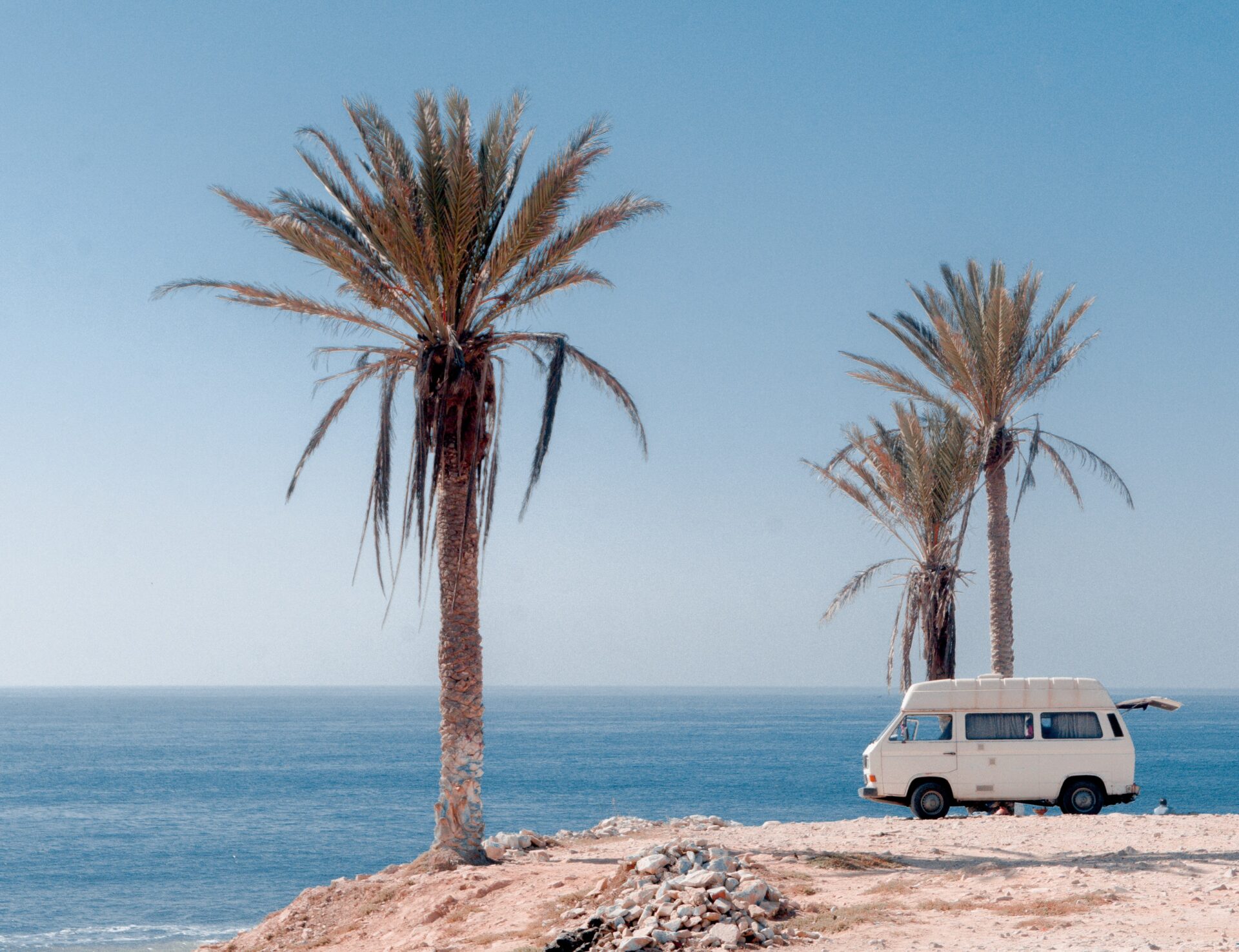 A white campervan standing on a cliff. Sea in the background, two palm trees. This symbolises Freedom.