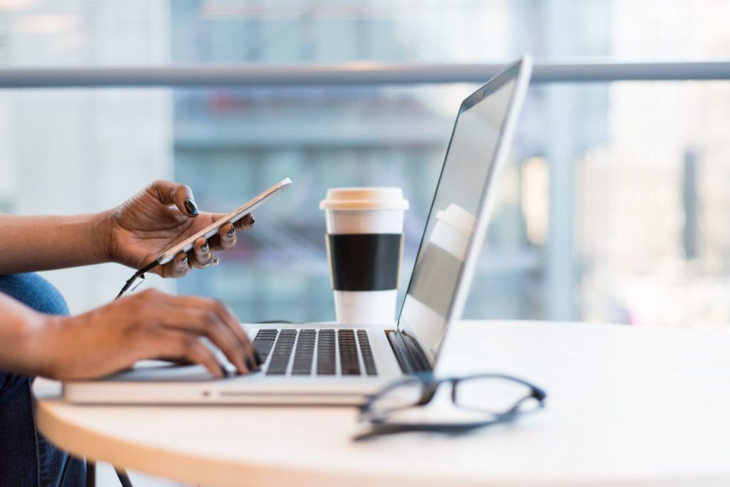 A laptop on a wooden desk. Next to it a coffee cup and two hands holding a phone.