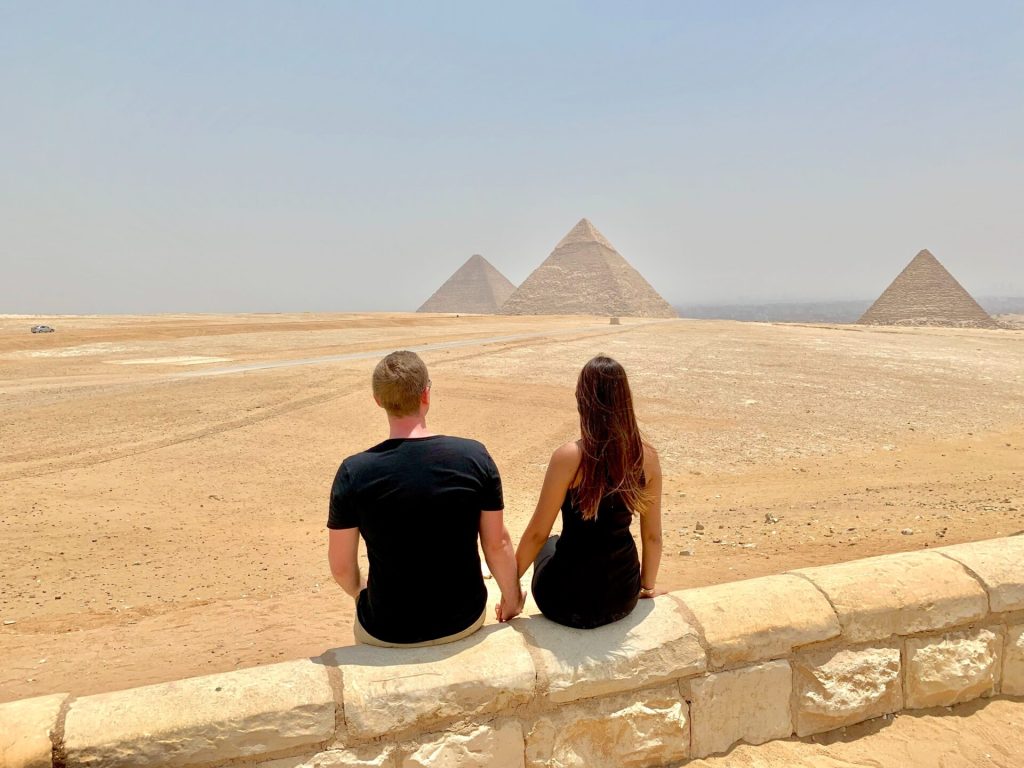 A young couple sitting on a wall in a desert watching two pyramids. Photo from behind.