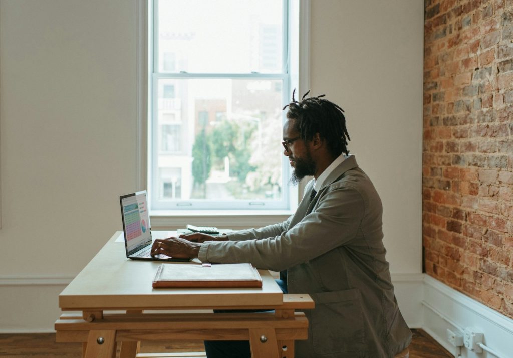 Afroamerican Man sitting on a wooden table working on his laptop next to a window