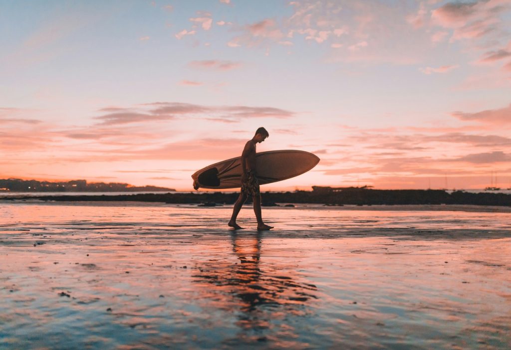 Man holding a surf board on a beach by sunset