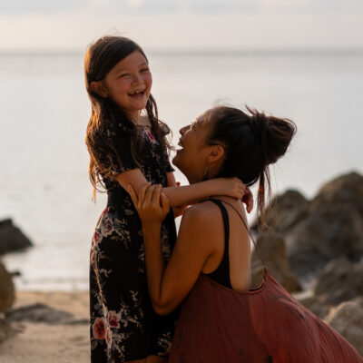 mother and daughter hugging each other on the beach, daughter laughs into camera, traveling as a single parent