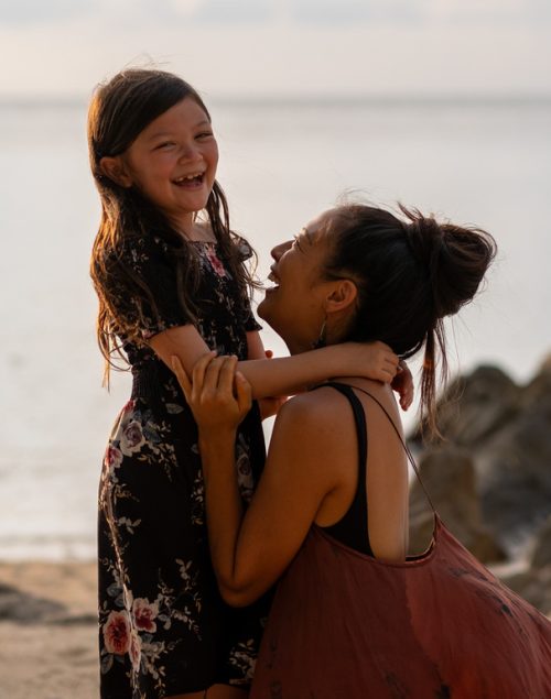 mother and daughter hugging each other on the beach, daughter laughs into camera, traveling as a single parent