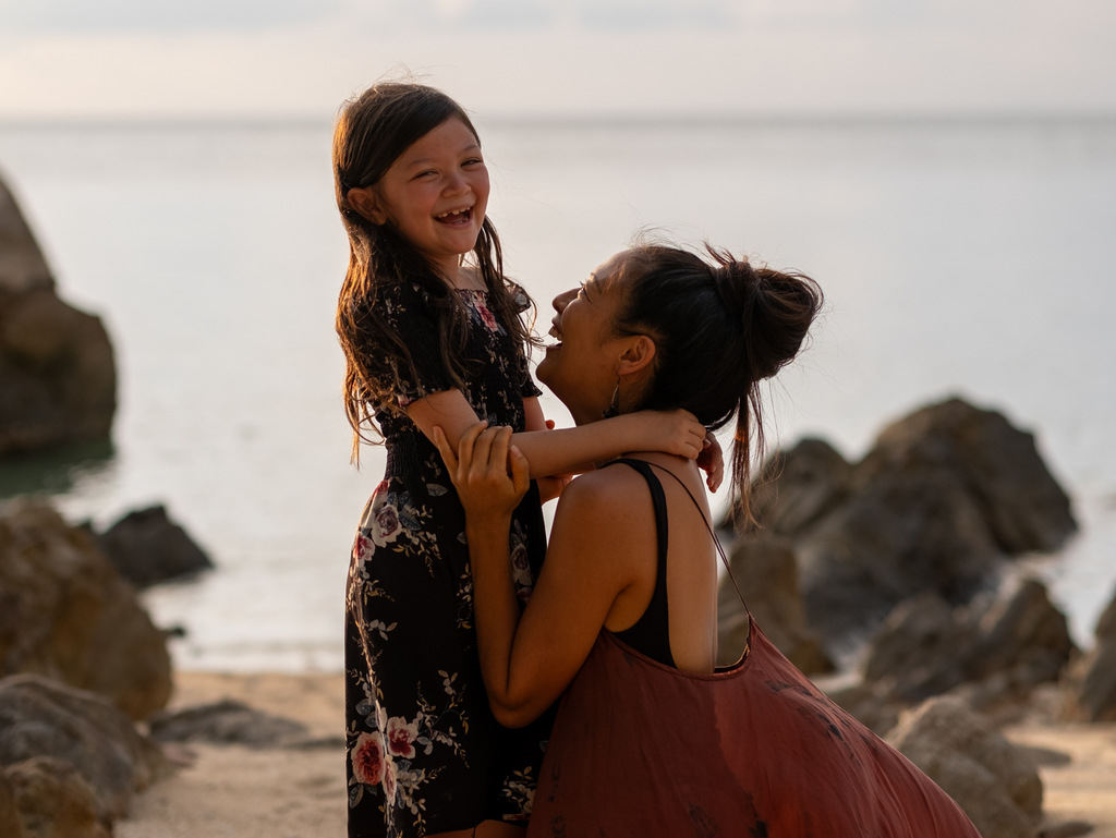 mother and daughter hugging each other on the beach, daughter laughs into camera, traveling as a single parent