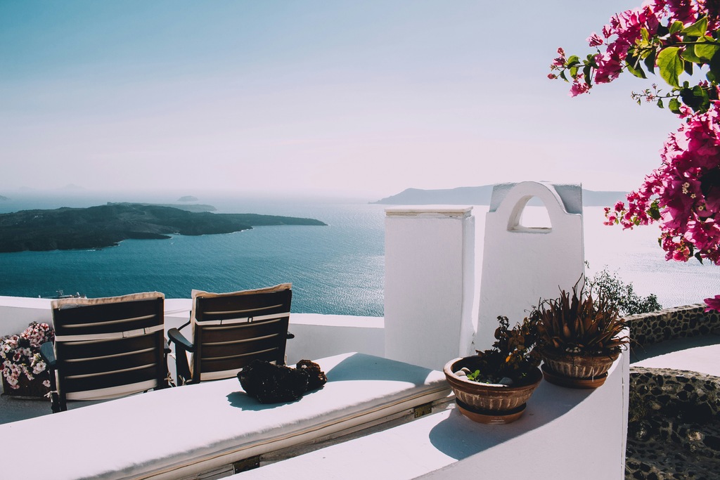 Two lounge chairs on a white terrace face the Aegean Sea, with vibrant pink bougainvillea on the right and potted plants in the foreground, capturing the tranquil beauty of a Greek island. Greece Digital Nomad Visa 2024