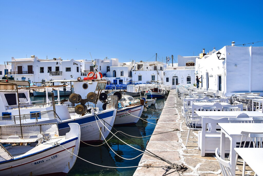 A picturesque harbor in Paros, Greece, featuring traditional fishing boats docked alongside white-washed buildings and outdoor café seating under a bright blue sky