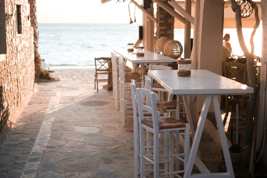 Seaside café in Naxos, Greece, featuring white tables and chairs, rustic stone walls, and a serene view of the ocean at sunset.