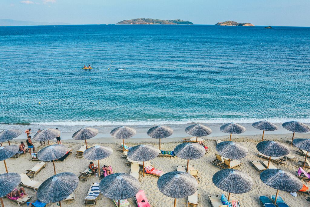 A scenic beach in Greece with rows of straw umbrellas and lounge chairs on the sandy shore, with the clear blue waters of the Aegean Sea and small islands in the background