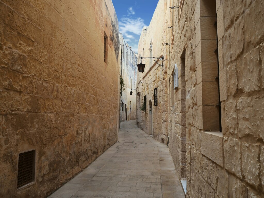 A narrow, quiet street in Mdina, Malta, lined with historic, beige stone buildings under a clear blue sky.