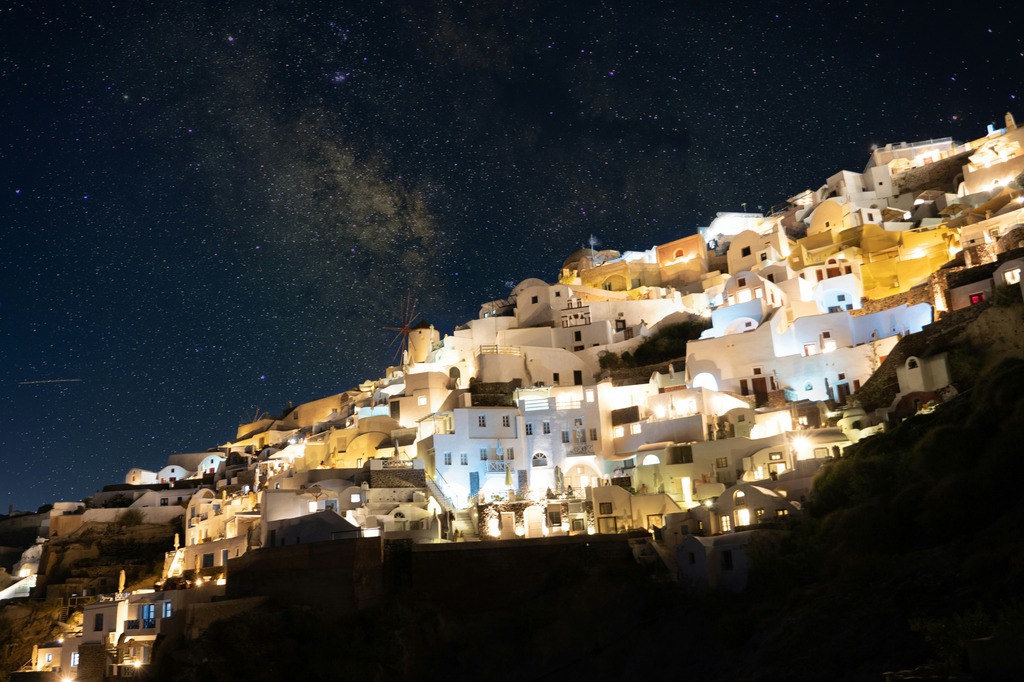 A night view of the village of Oia in Santorini, Greece, with its white-washed buildings illuminated against a backdrop of a starry sky, creating a magical and serene atmosphere.