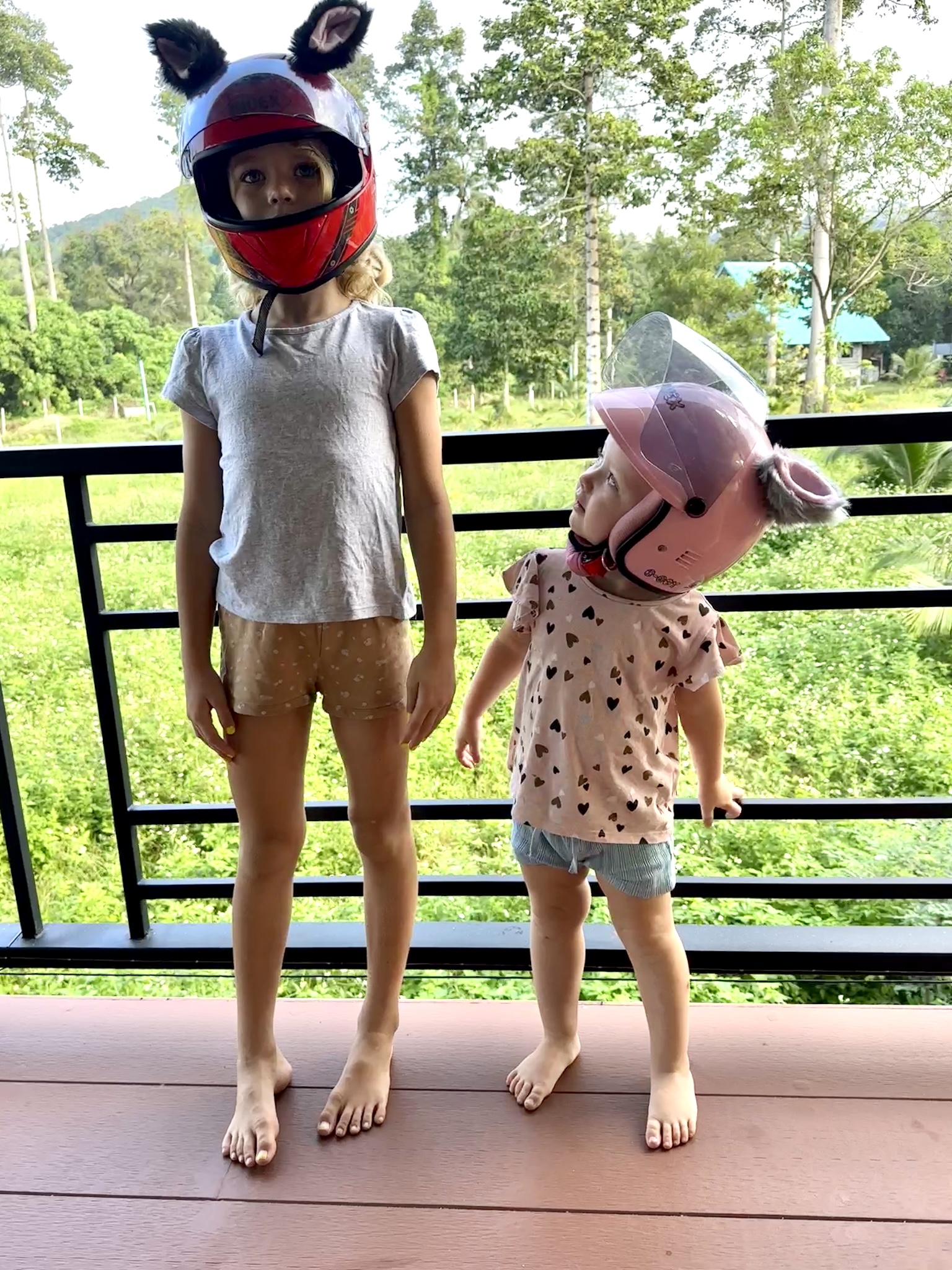 Two young children are standing on a porch, both wearing helmets with animal ears attached. The older child is wearing a red helmet with black ears, while the younger child is looking up at the older child, wearing a pink helmet with gray ears. They are both barefoot, and a lush green landscape is visible in the background.