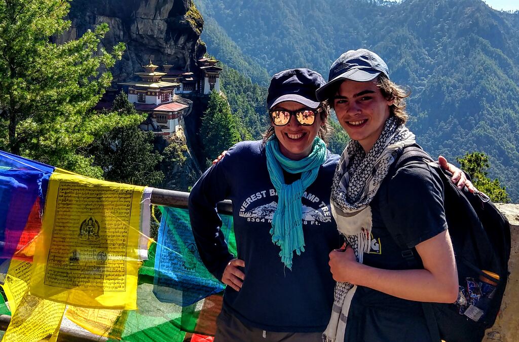 A mother and her teenage son are standing in front of the Tiger's Nest Monastery in Bhutan. They are both wearing hats, sunglasses, and scarves, smiling happily. 