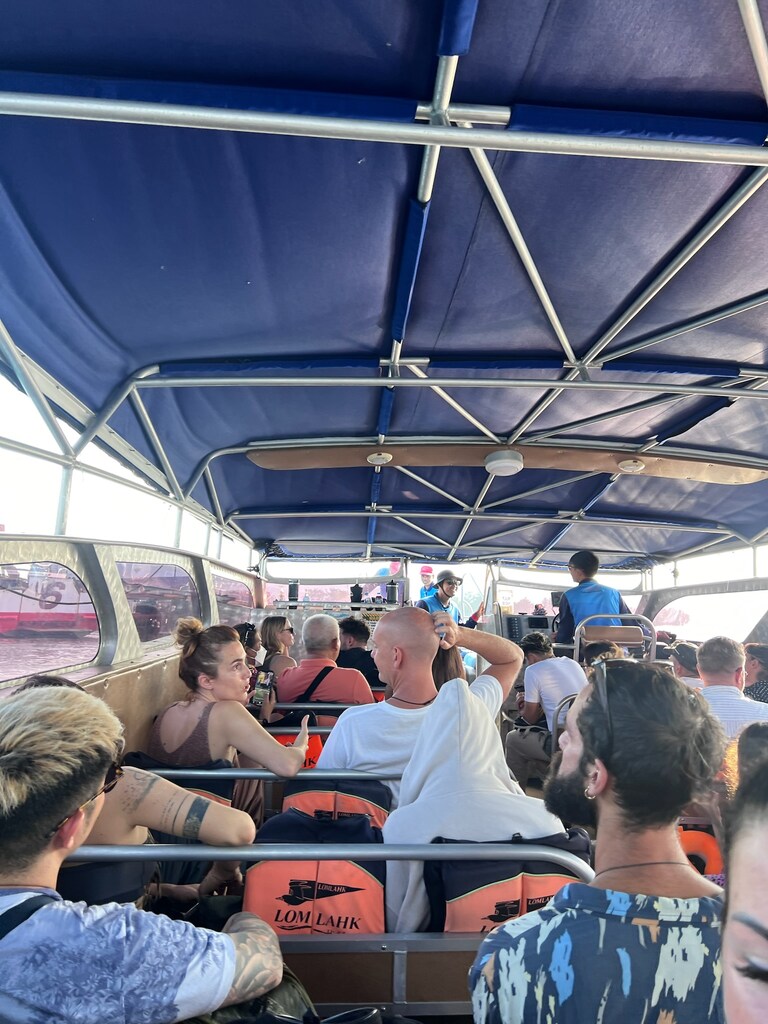 Passengers seated inside a covered boat, wearing life jackets and engaged in conversation. The boat's interior features blue fabric covering and metal frame structures. The atmosphere is casual, with people relaxing during the journey.