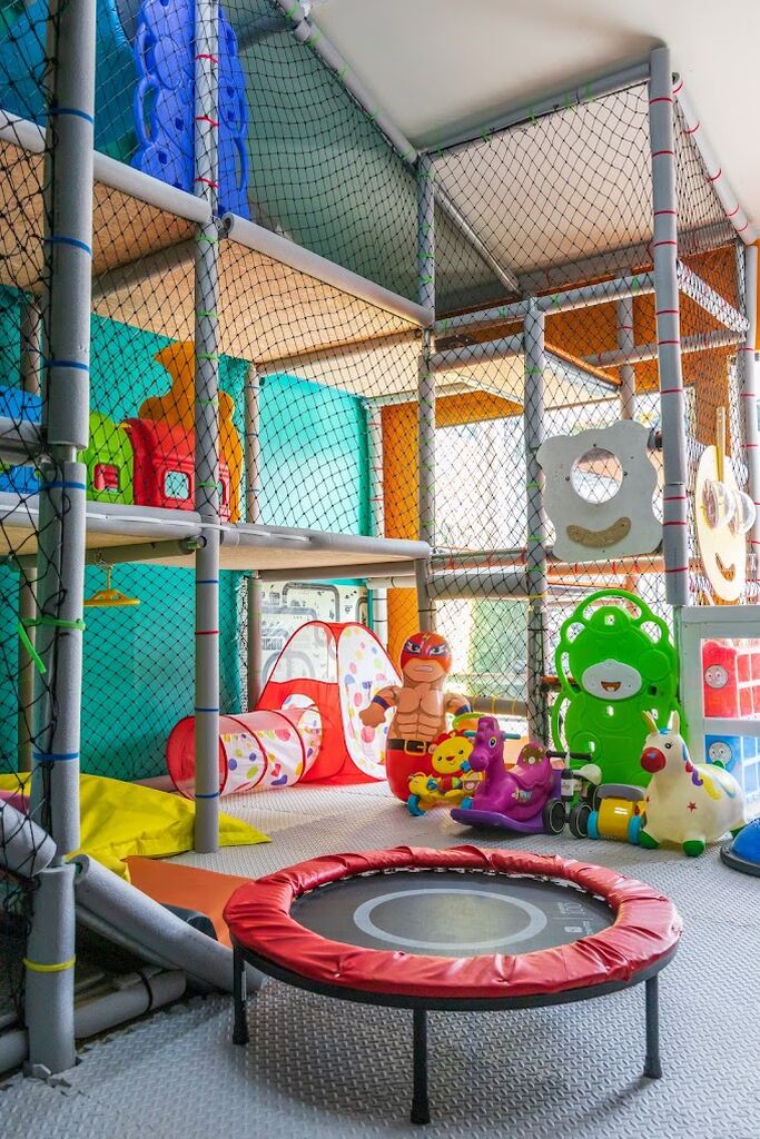 A colorful indoor play area for children featuring a soft trampoline, climbing structures, tunnels, and various playful toys including a rocking horse, a cartoonish muscleman figure, and a caterpillar-shaped climbing frame. The space is enclosed with safety nets and has a bright, cheerful ambiance, making it a fun and secure environment for kids.