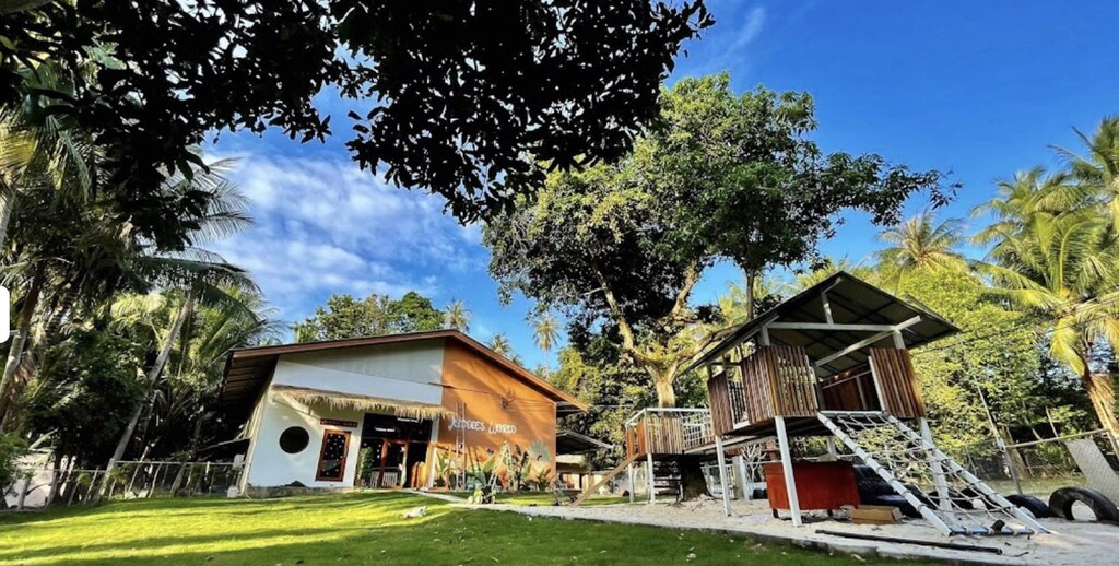 Vibrant and welcoming outdoor space at Kiddies World Nursery & Kindergarten. The scene features a well-maintained green lawn, a wooden playground structure with a climbing net and slide, and a building with large windows that likely serves as the main facility.