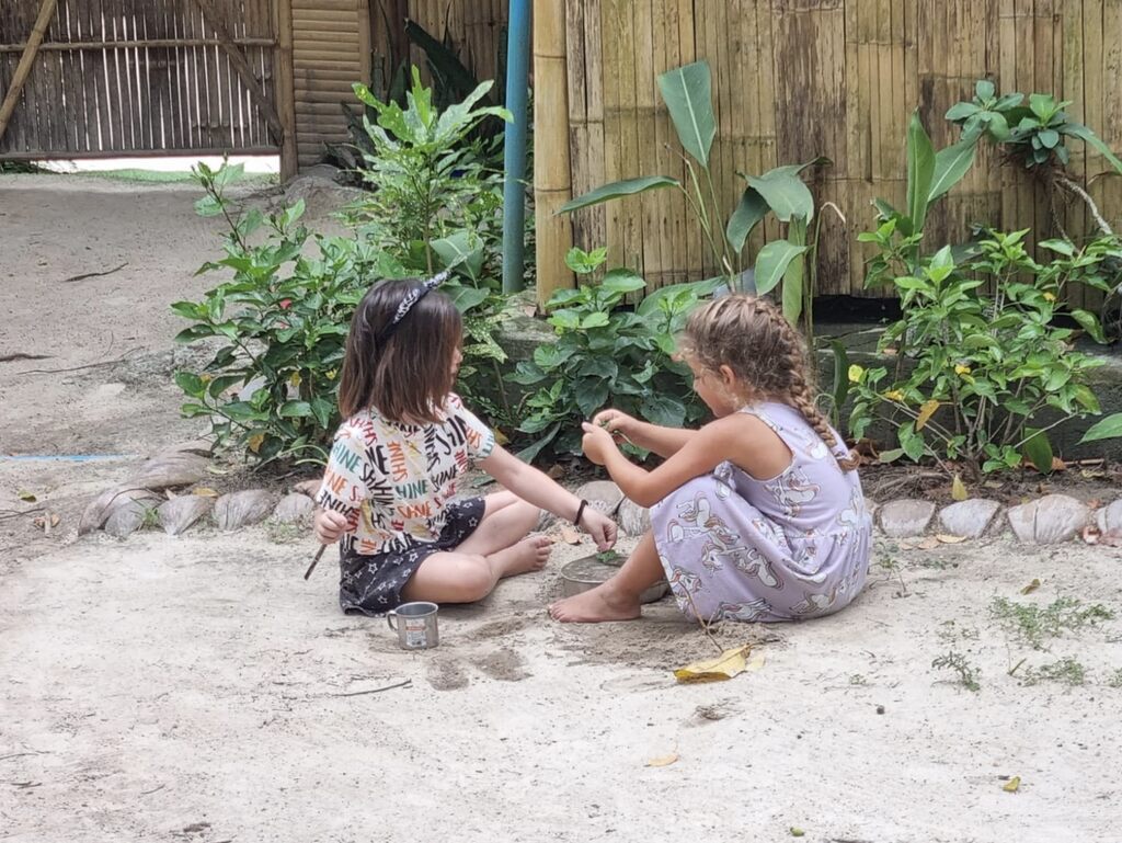 Two young girls are sitting barefoot on the ground outside, surrounded by greenery and bamboo structures.