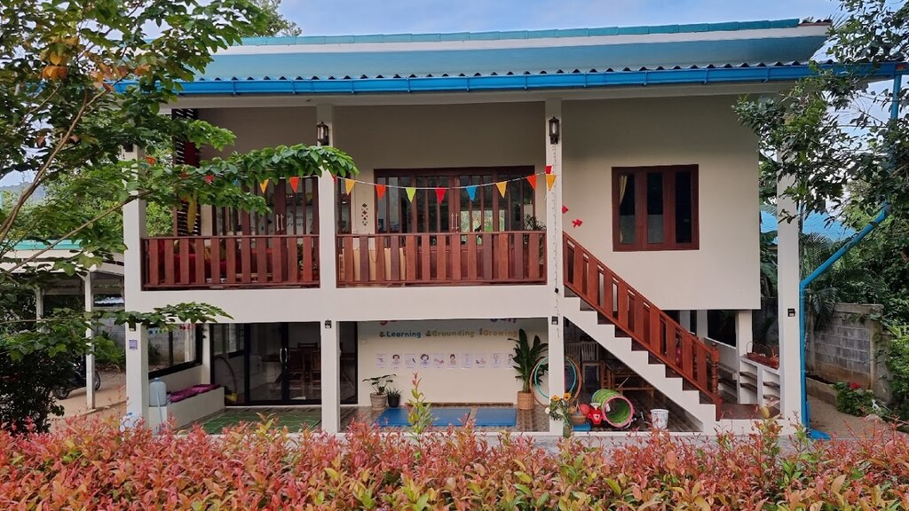 A two-story building with a simple and clean design. The exterior walls are painted white, and the roof is blue. The upper floor features a wooden balcony with a red railing and a small staircase leading to the ground level.