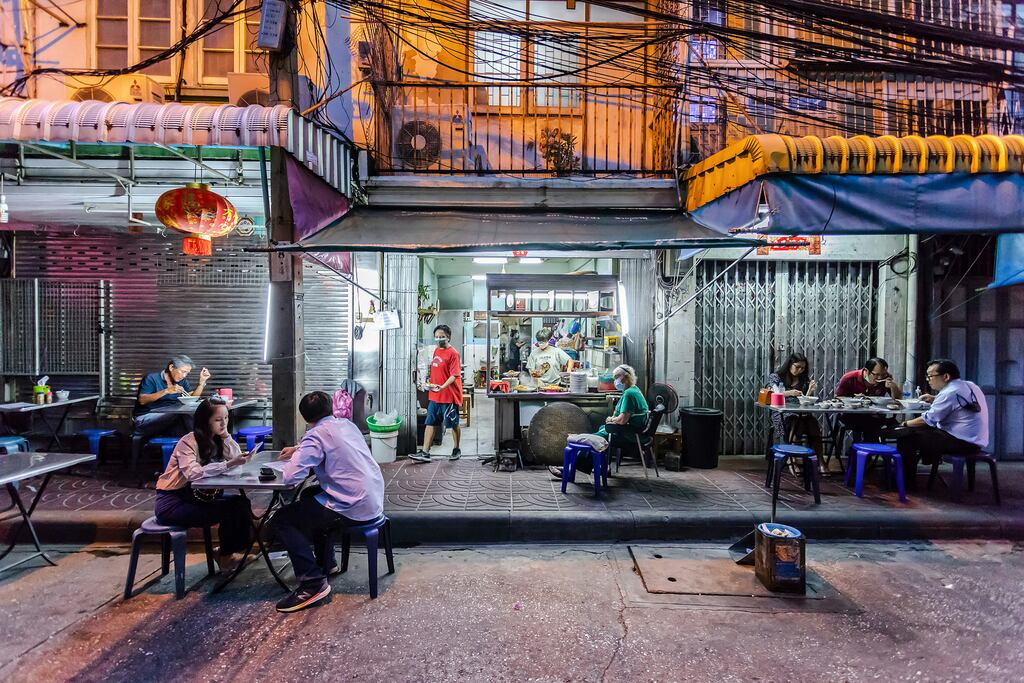 a lively street food scene in Thailand, where customers are seated at small tables along the sidewalk, enjoying their meals