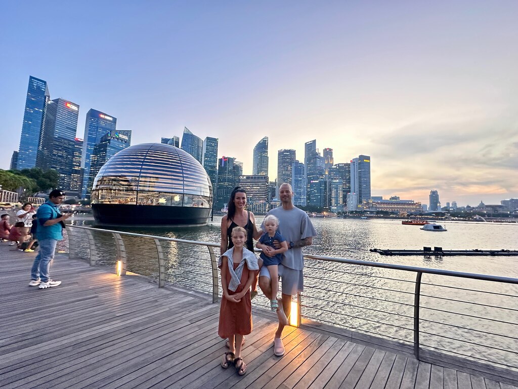 A family of four stands on a wooden boardwalk at Marina Bay, Singapore, with the iconic Apple store dome and a city skyline illuminated at dusk in the background, singapore with kids itinerary.