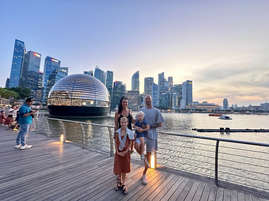 A family of four stands on a wooden boardwalk at Marina Bay, Singapore, with the iconic Apple store dome and a city skyline illuminated at dusk in the background, singapore with kids itinerary