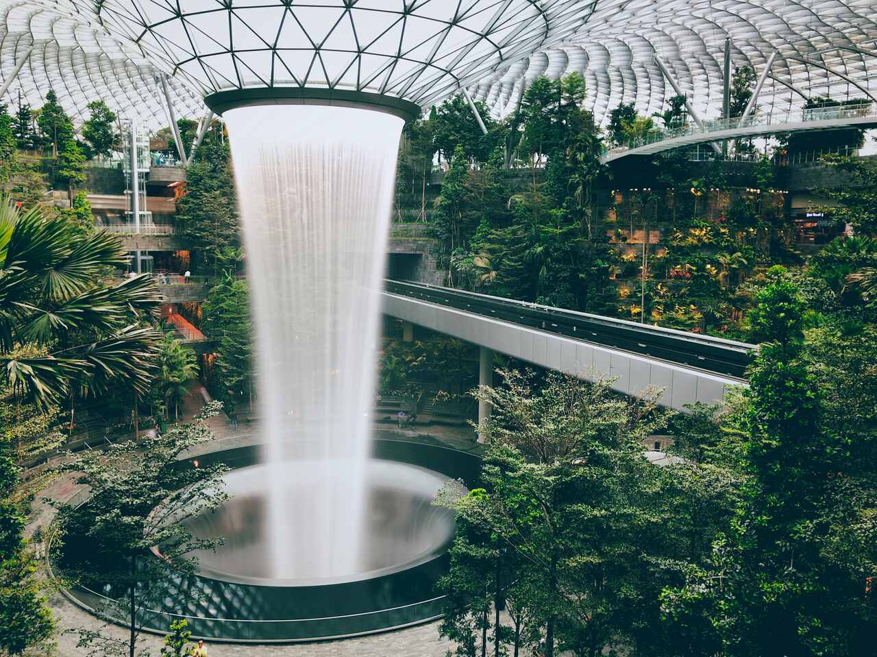 The Rain Vortex at Jewel Changi Airport, a massive indoor waterfall cascading into a circular basin, surrounded by greenery and modern architectural elements within a glass-domed structure.