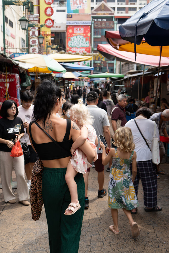 Mother carrying a young child while walking through a crowded street market in Kuala Lumpur, with her older daughter walking ahead.