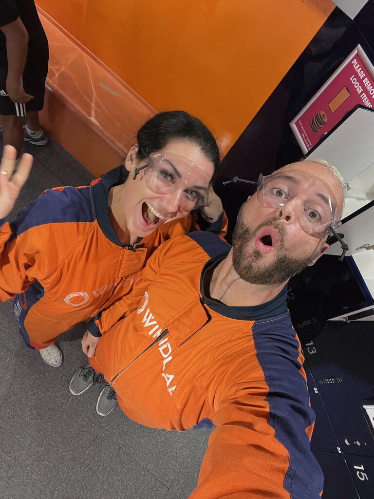 A man and woman in bright orange Windlab suits, wearing protective goggles, smiling and making excited faces before their indoor skydiving session at Windlab in Kuala Lumpur.