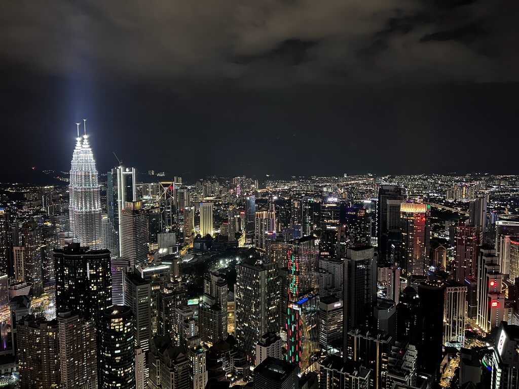 A nighttime view of Kuala Lumpur’s skyline, with the brightly lit Petronas Twin Towers standing out among numerous skyscrapers, all glowing under the dark sky.