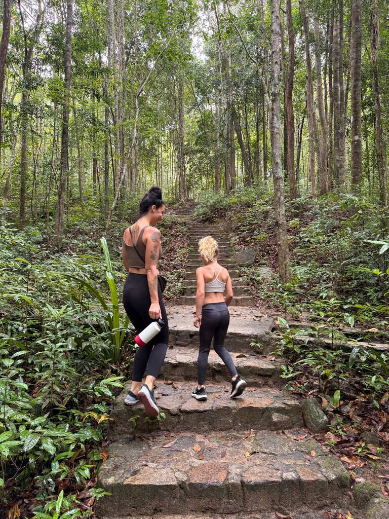 Mother and daughter from behind, walking on a jungle path, holding water bottles.