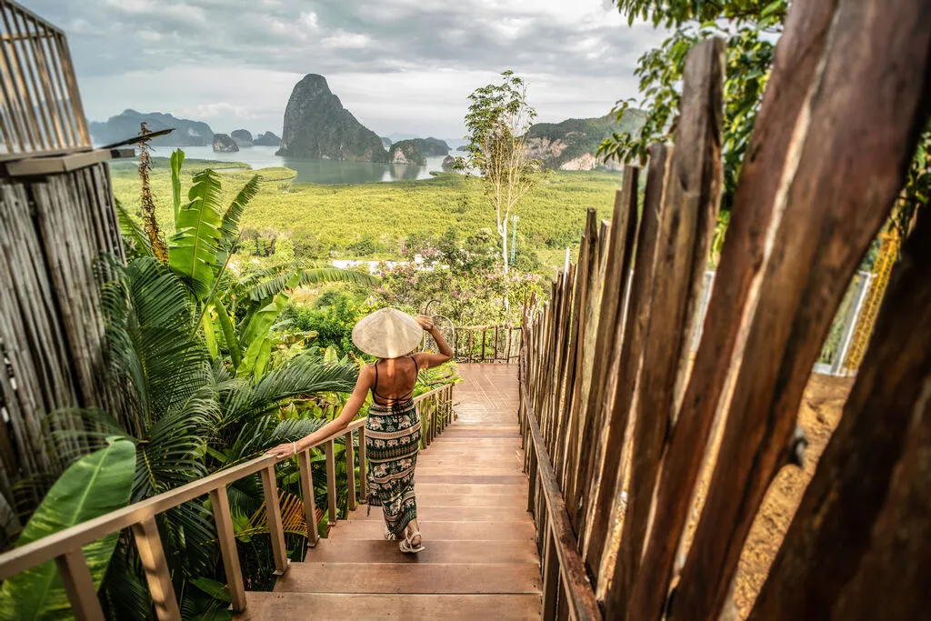 Woman with elephant dress and bamboo hat walking down a wooden path with beautiful lush landscape in the background. What to wear in Thailand.