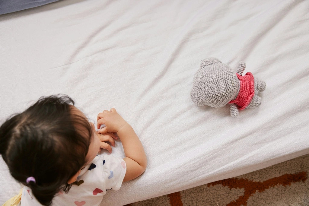 Black-haired toddler girl laying on a matress with white bed sheet.