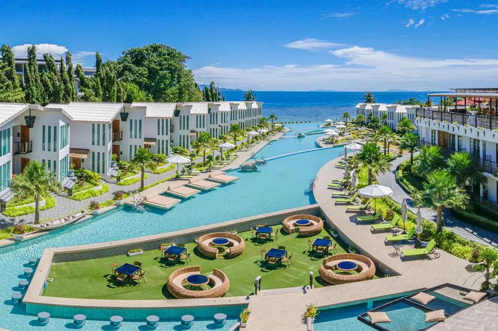 Aerial view of large hotel pool by the sea, surrounded by modern white villas left and right.