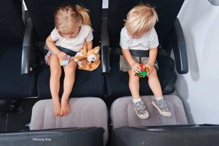 A young boy and girl sitting next to each other on a plane, both resting their feet on inflatable cushions. Airplane beds for toddlers and babies