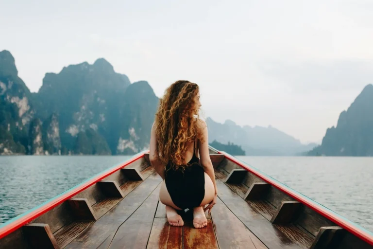 Woman with black swimsuit sitting on wooden boat with hills in the background. Best months to visit Thailand.