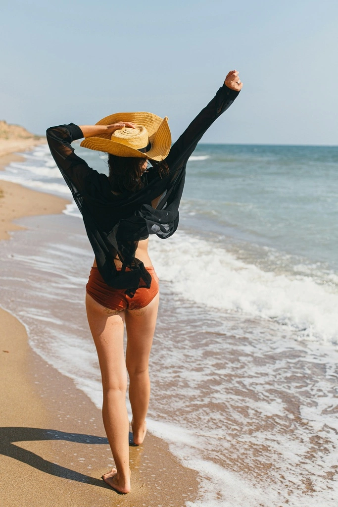 Woman on the beach wearing a bikini, a black blouse and a hat.