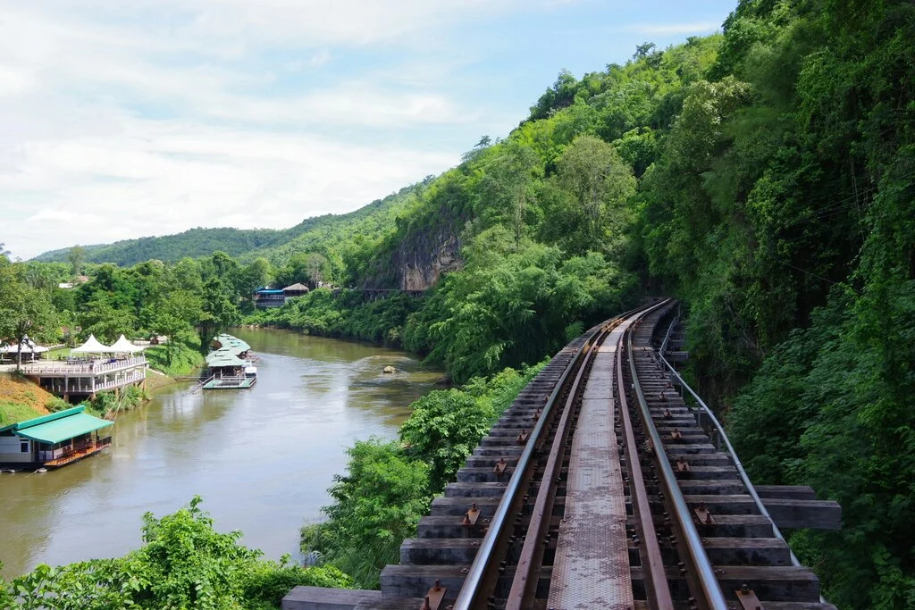 A lush, green riverside scene with floating houses and a railway track running along a cliffside.