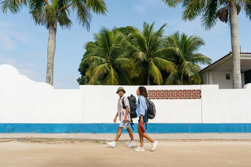 Young couple with backpacks walking by a white wall with palm trees in the background.