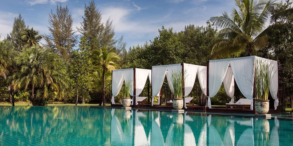 Huge pool with wooden sunbeds and white curtains, surrounded by trees.