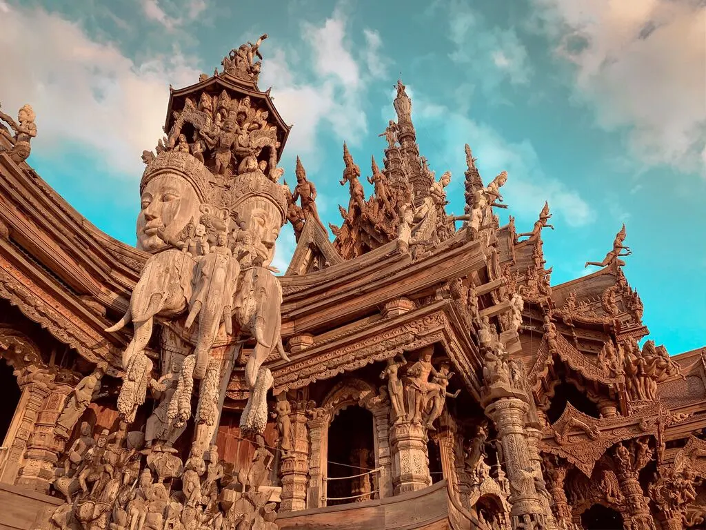 Intricately carved wooden facade of the Sanctuary of Truth in Pattaya, Thailand, featuring detailed sculptures of deities, elephants, and mythological figures against a blue sky.