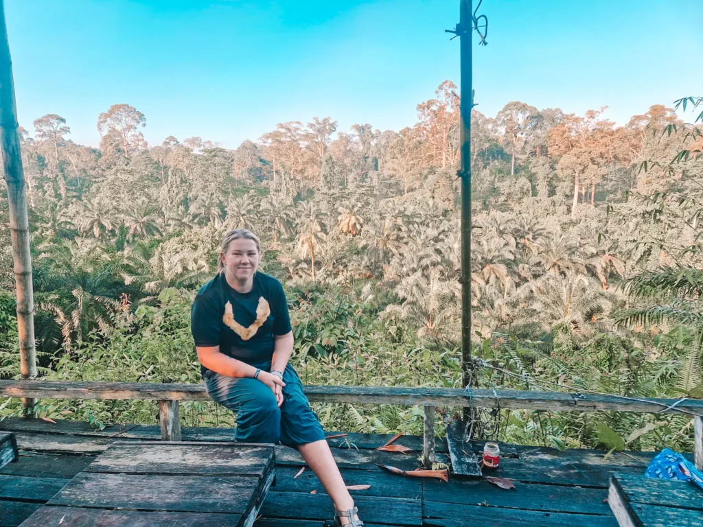 Epic 5-Day Borneo Itinerary: Meet Orangutans & Sun Bears 14 A person with light skin and blonde hair sits on a rustic wooden platform, wearing a black t-shirt with a sun bear crest design and blue pants. Behind them, an expansive jungle with tall trees and thick palm foliage stretches under a clear blue sky.