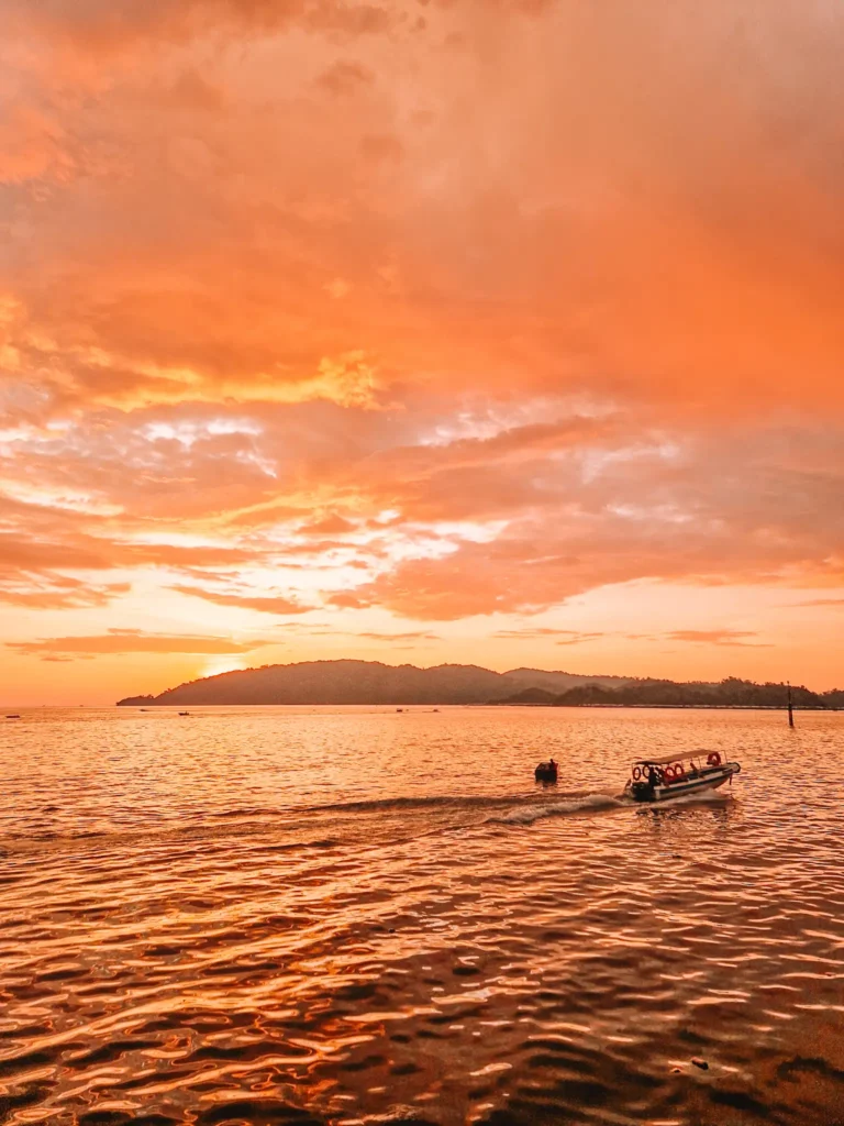 Epic 5-Day Borneo Itinerary: Meet Orangutans & Sun Bears 13 A breathtaking sunset with golden-orange hues reflecting off the rippling water. A small motorboat leaves a gentle wake as it moves toward the horizon, with distant hills silhouetted against the glowing sky.
