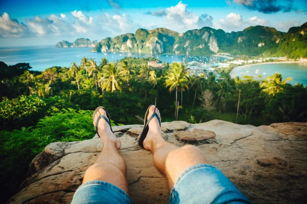 A person's legs and feet on a cliff overlooking a tropical island.