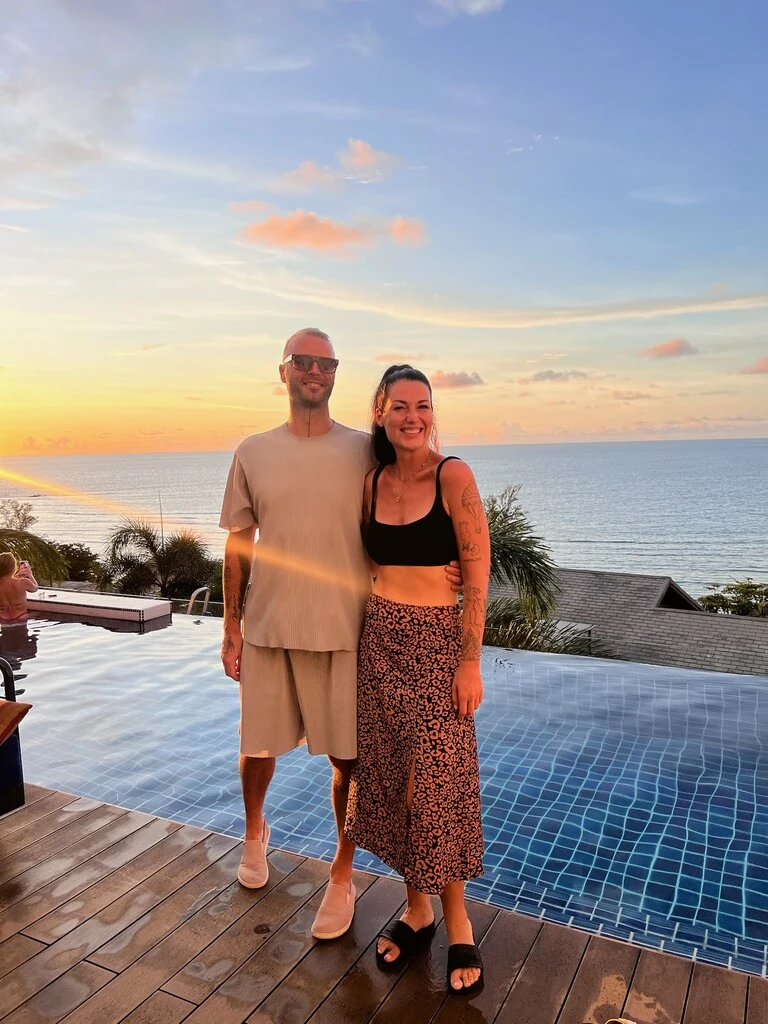 Hyatt Regency Phuket with Kids – My Honest Review 2 A man and woman standing next to a pool with a sunset in the background.