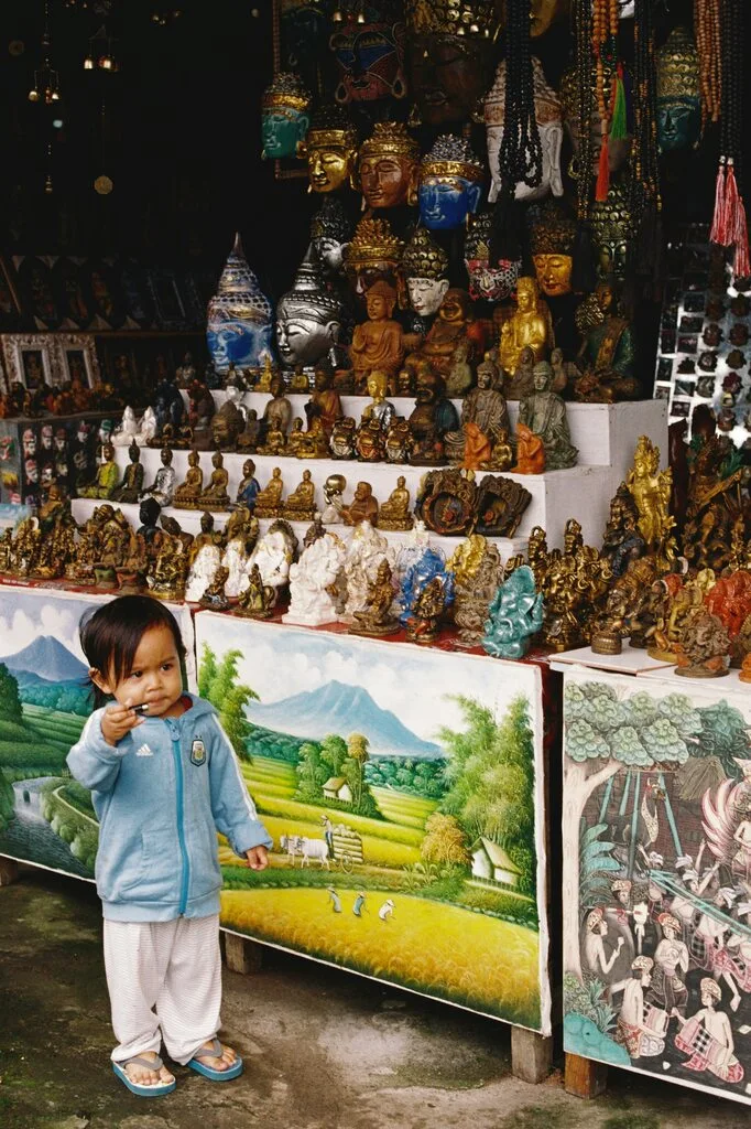 A young kid standing in front of a street stall will loads of buddha statues.