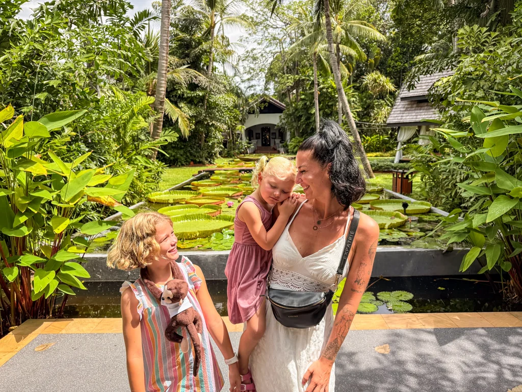 A woman and two girls standing on a walkway with water lilies and trees.