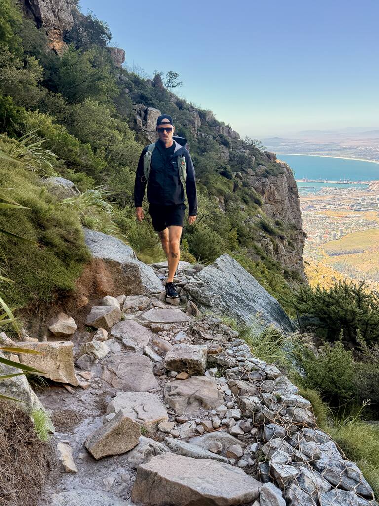 ChatGPT said: Man hiking along a rocky trail on Table Mountain with Cape Town city and the ocean in the background.